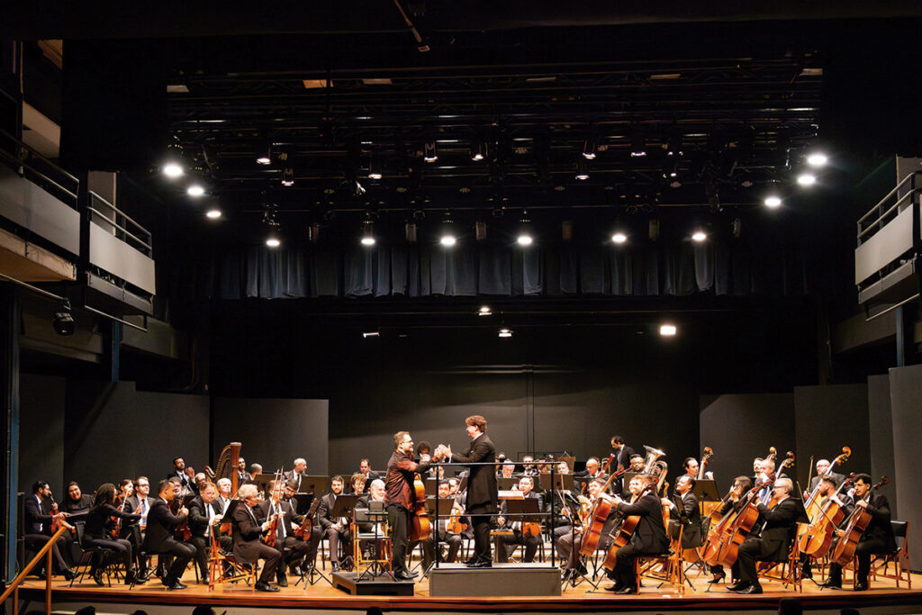 Orquestra sinfônica se apresenta em palco de teatro, com maestro ao centro e músicos tocando diversos instrumentos de corda e sopro.