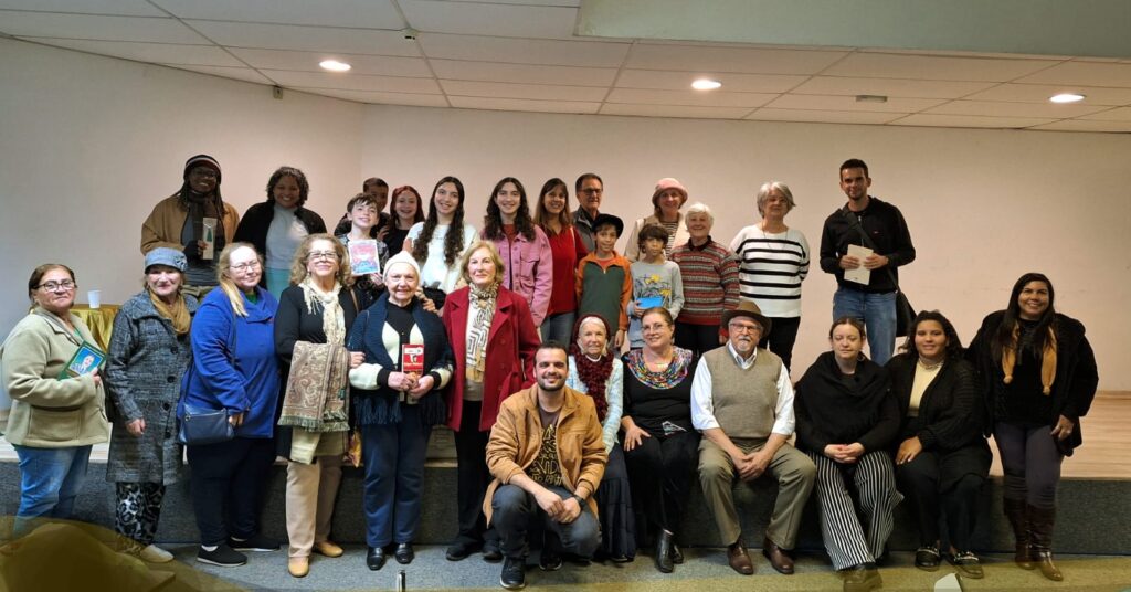 Cerca de 35 pessoas de diferentes idades posam sorrindo para a câmera, divididas entre pé e sentadas sobre o palco da Biblioteca Municipal. No grupo, há mulheres, homens, jovens, idosos e crianças. Algumas pessoas seguram livros. O cenário é simples, com fundo branco e teto de forro, indicando uma atividade cultural de integração.