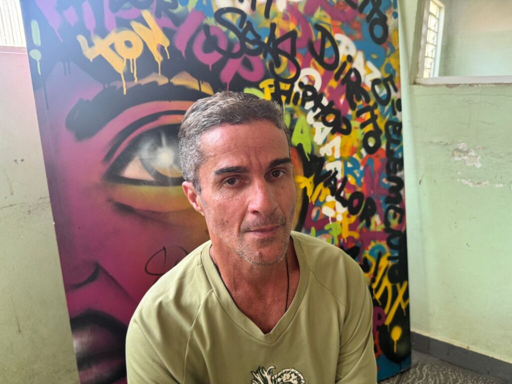 A imagem mostra um homem de meia-idade, com cabelos grisalhos e pele clara, olhando diretamente para a câmera. Ele está vestindo uma camiseta verde-oliva de gola V. Ao fundo, há uma grande obra de arte vibrante em estilo graffiti, dominada por cores como rosa, amarelo, azul e verde. A pintura parece representar um olho estilizado e várias palavras em letras grandes e coloridas, incluindo "HIP HOP" e "VALOR". A obra de arte está apoiada contra uma parede verde clara. O lado direito da imagem mostra parte de uma janela e o canto de uma sala.