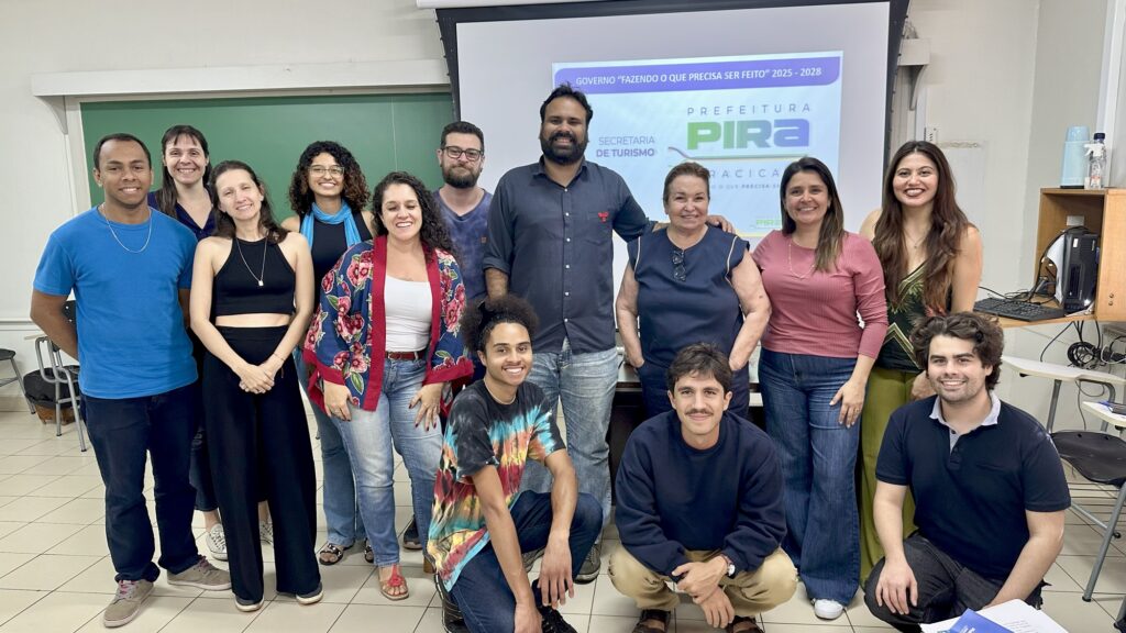 A imagem mostra um grupo de 12 pessoas em uma sala, possivelmente uma sala de aula ou de reunião. Elas estão posadas em frente a uma tela de projeção que exibe o texto "GOVERNO 'FAZENDO O QUE PRECISA SER FEITO' 2025-2028" e o logotipo da "PREFEITURA PIRA RACICA".As pessoas estão vestidas de forma casual e variam em idade e etnia. Algumas estão em pé e outras agachadas na frente. Todos sorriem para a câmera, sugerindo um ambiente positivo e colaborativo.A iluminação na sala parece ser artificial, vinda de cima. Há uma lousa verde ao fundo, à esquerda, e à direita, uma estante com equipamentos eletrônicos, como um computador e um monitor. O piso é de ladrilhos claros.No geral, a imagem transmite uma sensação de equipe ou grupo de trabalho reunido para uma apresentação ou evento relacionado à gestão pública, especificamente à Secretaria de Turismo da Prefeitura de Piracicaba.