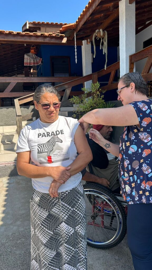 Na imagem, uma pessoa está recebendo uma injeção no braço esquerdo, aplicada por outra pessoa vestida com uma camisa azul estampada com personagens de desenho e itens esportivos. Quem recebe a injeção usa uma camiseta branca com a estampa de uma zebra e a palavra "PARADE", além de uma saia longa com padrão colorido. Ao fundo, vê-se uma pessoa em cadeira de rodas, uma mureta de madeira e uma casa com telhado de cerâmica e paredes azuis, sugerindo que a cena ocorre em um espaço externo, como um quintal ou pátio.
