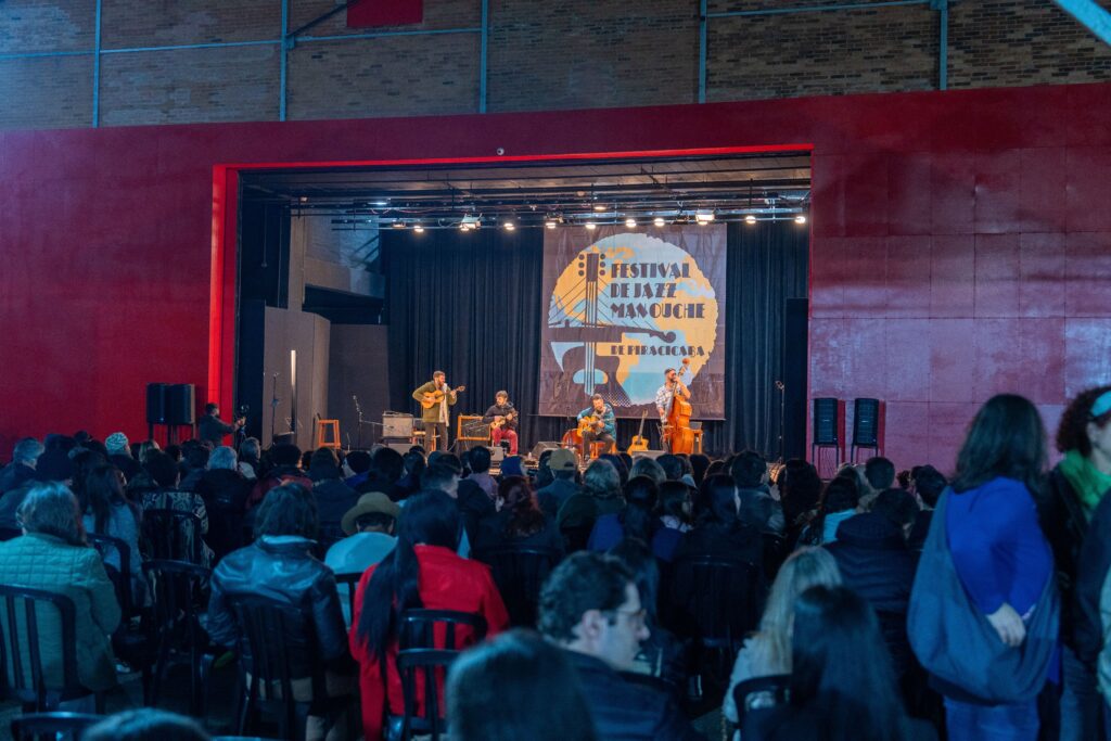 Foto mostra apresentação noturna no palco do Teatro do Engenho. Quatro músicos tocam guitarra e contrabaixo acústico sob iluminação amarelada. O painel de fundo traz a identidade visual do Festival de Jazz Manouche de Piracicaba. A plateia ocupa quase toda a área, com pessoas usando casacos e agasalhos devido ao clima frio.