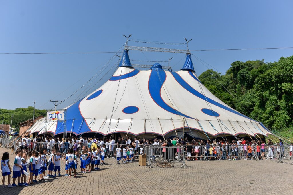 Vista externa de uma grande lona de circo branca com detalhes azuis. Ao redor, filas organizadas de crianças em uniforme escolar e muitos visitantes aguardam a entrada. O céu está limpo e azul, reforçando o clima de dia ensolarado.