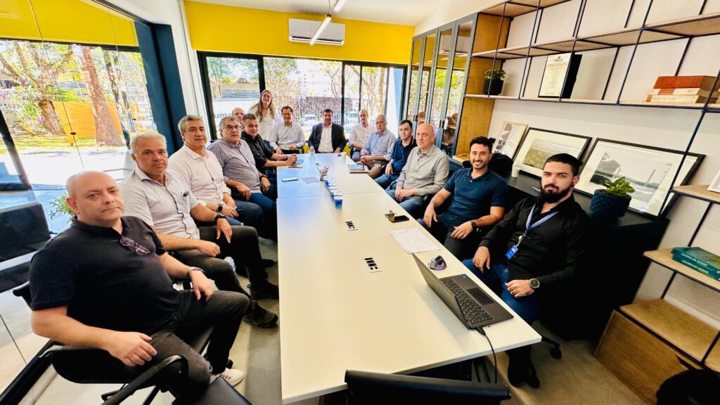 A imagem mostra um grupo de pessoas sentadas ao redor de uma mesa de conferência em uma sala moderna e bem iluminada. No centro da sala, há uma grande tela exibindo um fluxograma. A sala possui estantes altas com portas de vidro em um lado, revelando prateleiras cheias de livros e objetos decorativos. Do outro lado, uma parede de vidro permite ver um escritório aberto com mesas de trabalho e cadeiras. As pessoas na mesa de conferência parecem estar engajadas em uma reunião, com alguns olhando para a tela, outros conversando entre si. A atmosfera geral sugere um ambiente de trabalho colaborativo e profissional.