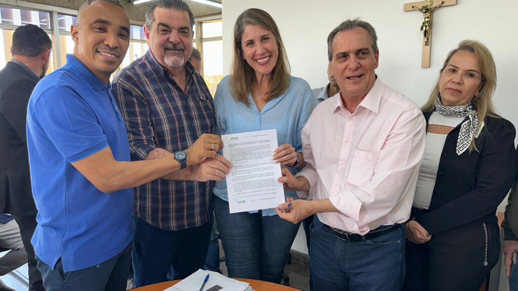 Quatro pessoas posam sorrindo para a foto enquanto seguram juntas o termo de permissão assinado. Uma mulher de camisa azul segura o documento ao centro. Ao fundo, a parede branca com um crucifixo pendurado.