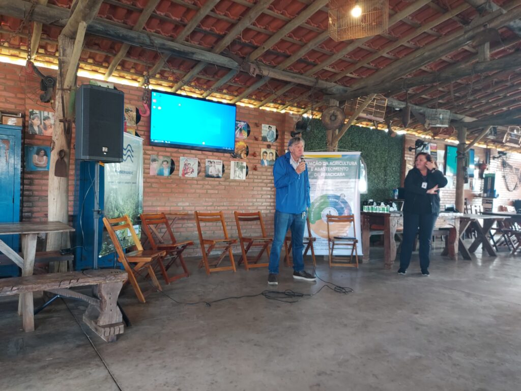 A imagem mostra um ambiente rústico com estrutura de madeira e telhado de telhas, preparado para receber um público. Cadeiras de madeira estão organizadas em fileiras voltadas para a frente, onde há um banner institucional da Secretaria de Agricultura e Abastecimento de Piracicaba, acompanhado de um gráfico circular. Duas pessoas estão em pé, uma delas com microfone, indicando que estão conduzindo uma apresentação ou palestra. Na parede, uma televisão grande exibe uma tela azul, sugerindo que uma apresentação digital está prestes a começar.