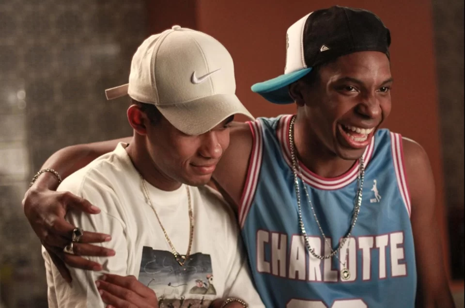 Dois jovens estão abraçados e demonstram amizade e alegria. O da esquerda veste boné branco da Nike, camiseta clara com estampa e correntes prateadas no pescoço. Ele sorri de forma contida e parece emocionado. O da direita veste regata azul clara de basquete com a palavra “Charlotte” em branco, boné preto e corrente prateada. Ele ri abertamente, transmitindo entusiasmo e felicidade.