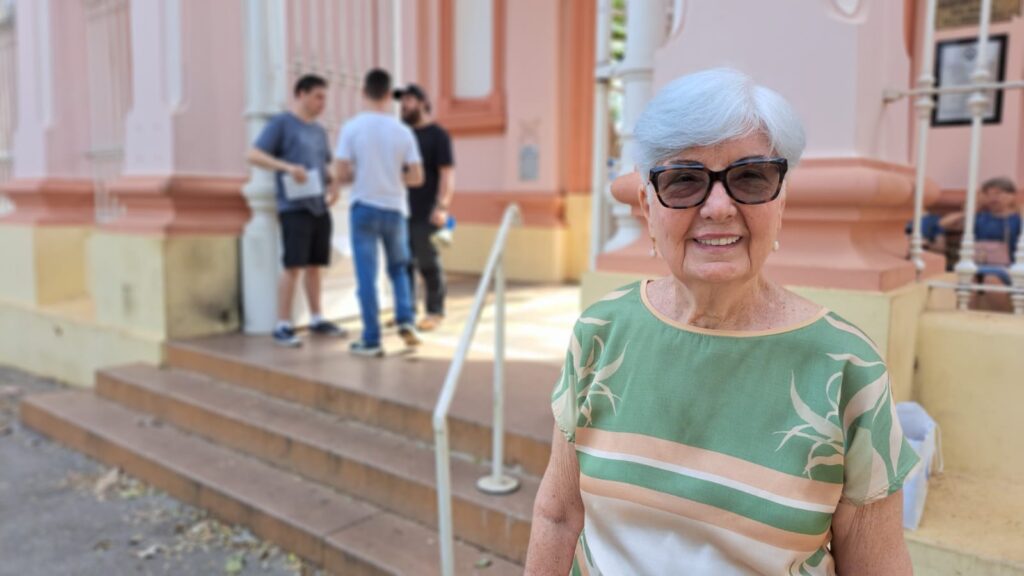 Uma senhora idosa sorri para a câmera, com o cabelo grisalho curto e óculos pretos. Ela usa uma blusa verde com um padrão de folhas e listras horizontais em tons de pêssego e verde claro.Ao fundo, desfocadas, estão algumas pessoas em frente a um edifício com paredes cor-de-rosa e detalhes em amarelo e laranja nas escadas e colunas. Há também uma grade branca e alguns degraus de escada.