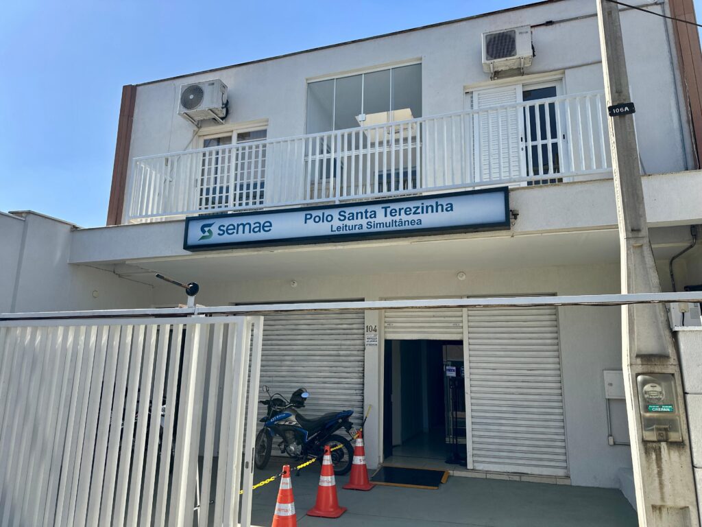 A imagem mostra a fachada de um edifício com a sinalização "SEMAE Polo Santa Terezinha Leitura Simultânea". O edifício tem uma varanda com grades brancas no andar superior, onde estão instalados dois aparelhos de ar condicionado. Abaixo da varanda, há uma placa iluminada com o nome do local. No térreo, há um portão branco, uma motocicleta estacionada e cones de sinalização. Uma porta de acesso principal está aberta, revelando o interior do estabelecimento. Há também uma caixa de medidor de energia elétrica presa a um poste na lateral direita da imagem. O céu está limpo e azul.