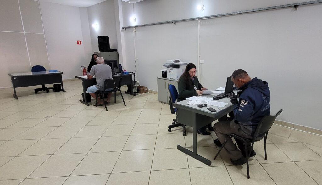 A imagem mostra um interior de um escritório com várias pessoas interagindo em balcões. No lado esquerdo, um homem com cabelo grisalho e camiseta cinza está sentado em uma cadeira preta em frente a um balcão escuro, com uma mulher com cabelo escuro sentada à sua frente. Há um extintor vermelho e um extintor de incêndio em pé no balcão. Outro balcão vazio está posicionado à esquerda. No lado direito, uma mulher com óculos e cabelo escuro está sentada em uma cadeira azul em frente a um balcão, olhando para os papéis em sua frente. Ao lado dela, uma impressora multifuncional está sobre um gabinete cinza. Um homem com uma jaqueta azul escura está sentado em uma cadeira preta em frente a ela, olhando para um telefone em sua mão. Ambos os balcões estão equipados com computadores e papéis. O chão é revestido com azulejos claros em um padrão de diamante. As paredes são brancas, com algumas linhas verticais e horizontais em relevo. Há duas luzes fluorescentes no teto.