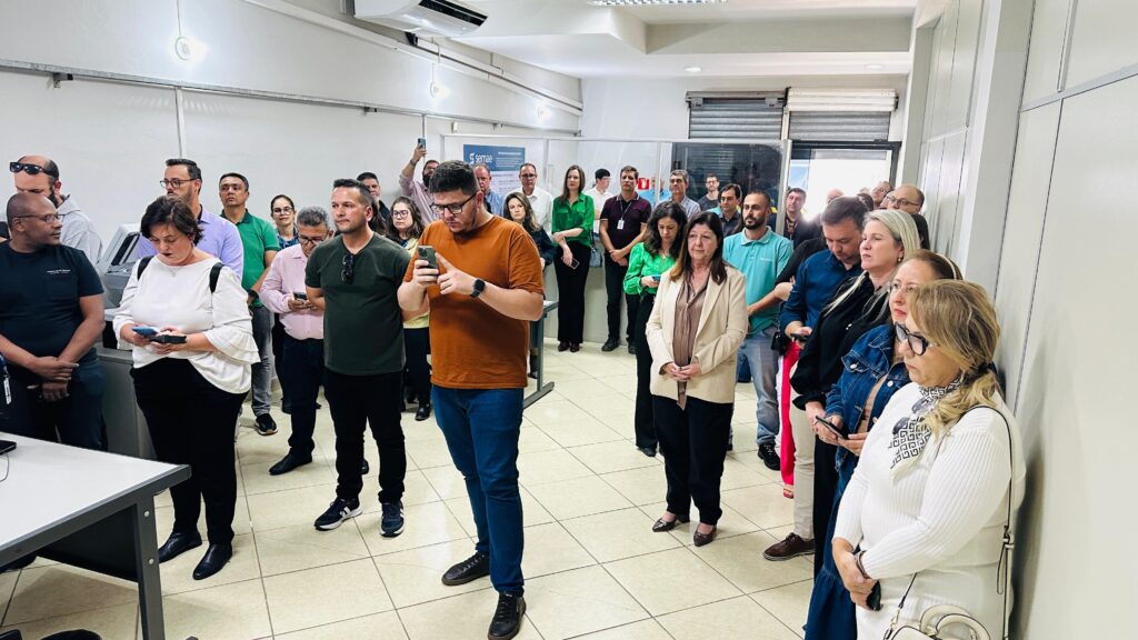 A imagem mostra um grupo de pessoas em pé, alinhadas em um corredor, possivelmente em um evento ou reunião. A maioria está olhando para a frente, e alguns parecem estar segurando celulares. Há uma mulher de blazer bege em primeiro plano, no lado direito da imagem, e um homem de camiseta laranja no lado esquerdo, também em primeiro plano, olhando para seu celular. Ao fundo, há uma placa com o logo "SEMAE". O ambiente parece ser um escritório ou uma sala de espera, com paredes brancas e piso quadriculado.