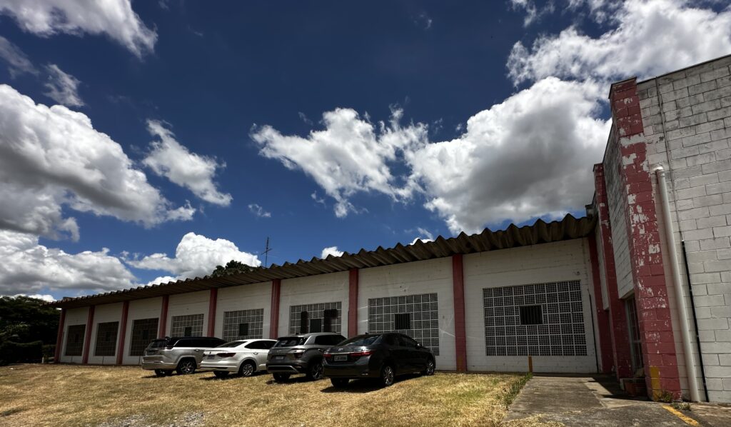 A imagem retrata um prédio com fachada de concreto e tijolos pintados em branco e vermelho, exibindo janelas gradeadas e um telhado ondulado. À frente da construção, há um estacionamento informal sobre grama seca, onde cinco carros de diferentes modelos e cores estão estacionados. O céu azul com nuvens esparsas indica um dia ensolarado.