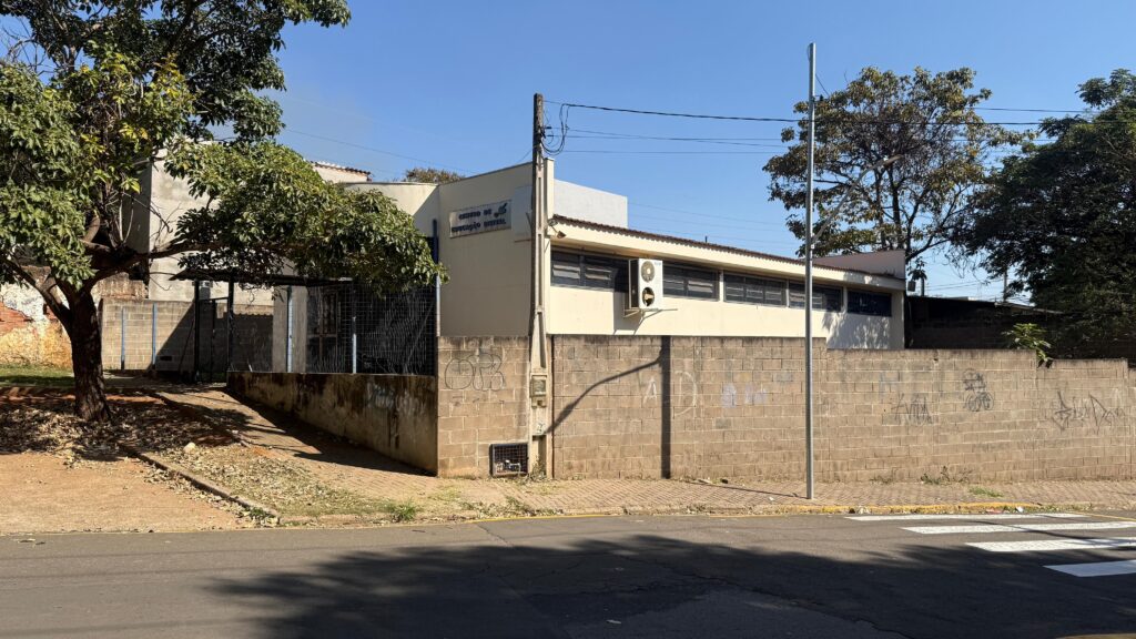 A imagem mostra a fachada de um edifício com a inscrição "CENTRO DE EDUCAÇÃO DIGITAL". O edifício é de cor creme e possui janelas com persianas. Há um ar condicionado instalado na parede externa. Na frente do edifício, há um muro de blocos de concreto com grafites. Uma árvore frondosa com folhas verdes está posicionada à esquerda da construção. O céu azul claro e a iluminação do sol indicam um dia ensolarado. Uma faixa de pedestres é visível na rua em primeiro plano.