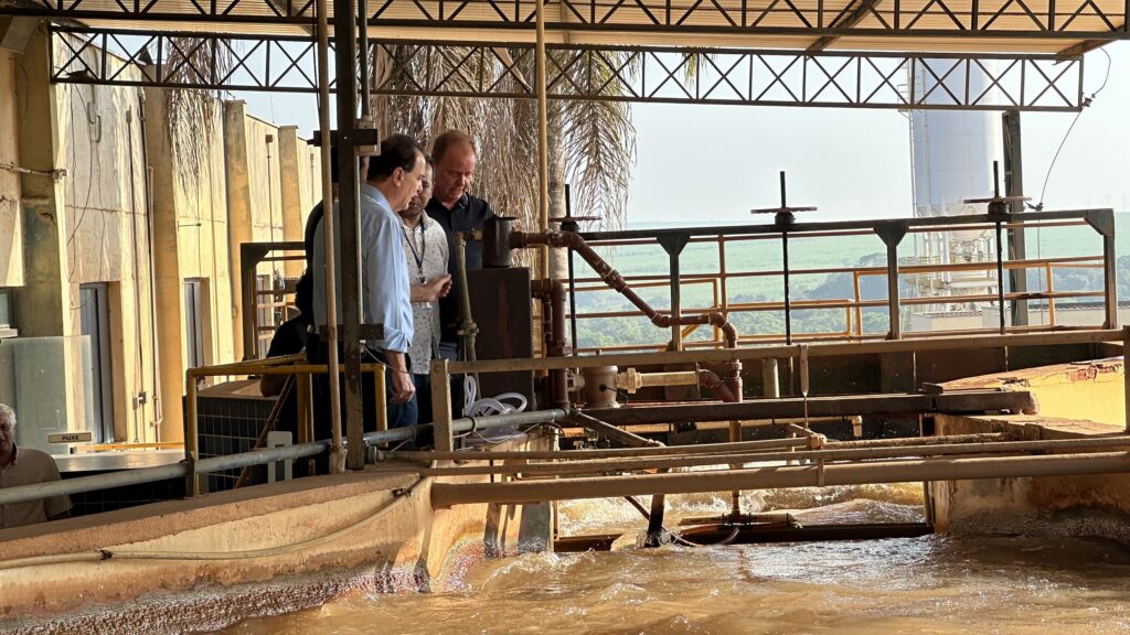 Três homens estão em um ambiente que parece ser industrial, possivelmente uma fábrica ou planta de processamento. Eles estão reunidos ao redor de algumas máquinas e tubos. O homem à esquerda veste uma camisa azul clara, o do meio usa uma camisa branca com estampa, e o homem à direita está com uma camisa escura. Ao fundo, há uma estrutura metálica com telhado, tubulações e, mais além, um campo verde e um silo branco alto. A cena passa a impressão de uma visita ou inspeção no local.