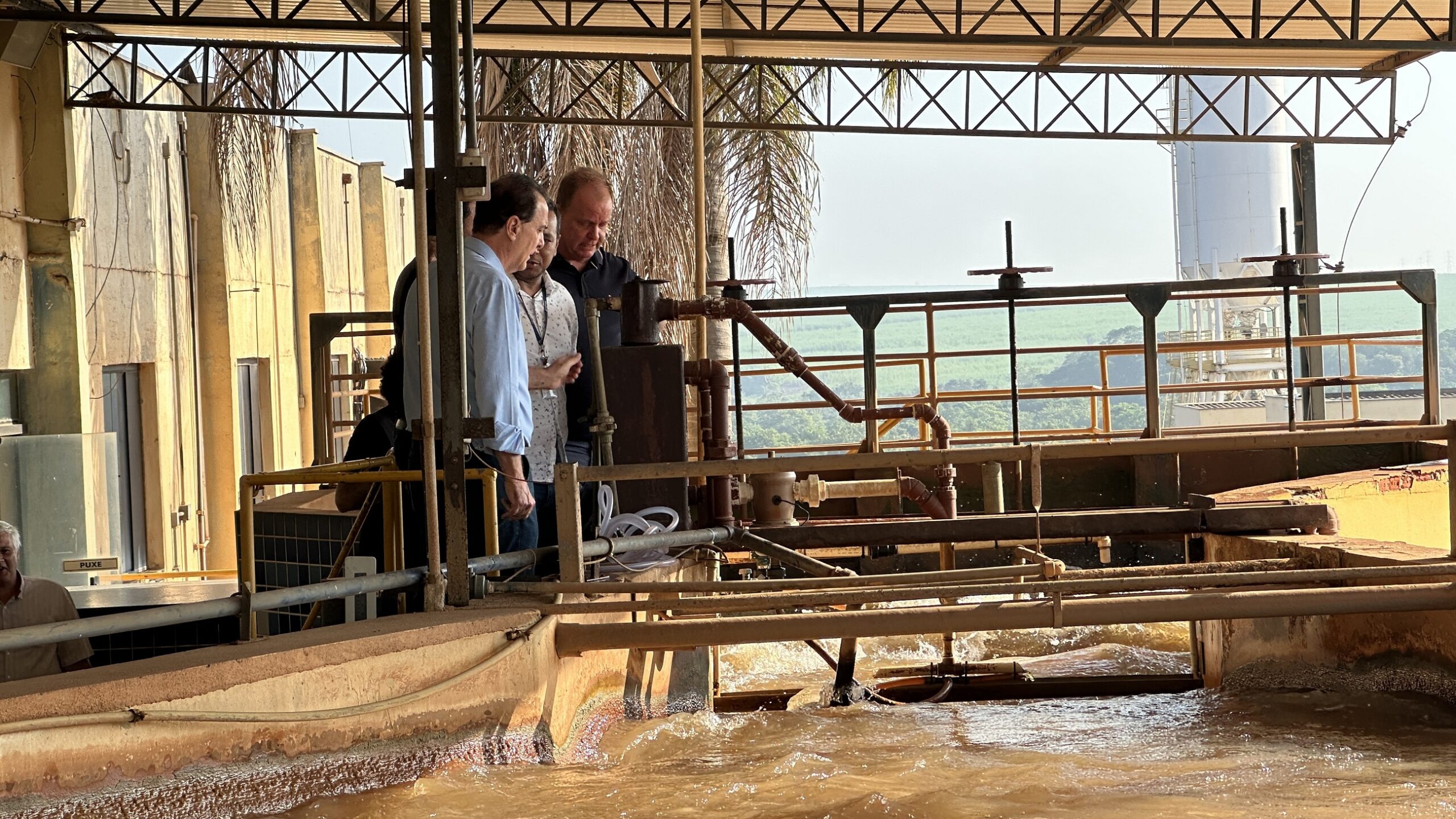 Três homens estão em um ambiente que parece ser industrial, possivelmente uma fábrica ou planta de processamento. Eles estão reunidos ao redor de algumas máquinas e tubos. O homem à esquerda veste uma camisa azul clara, o do meio usa uma camisa branca com estampa, e o homem à direita está com uma camisa escura. Ao fundo, há uma estrutura metálica com telhado, tubulações e, mais além, um campo verde e um silo branco alto. A cena passa a impressão de uma visita ou inspeção no local.
