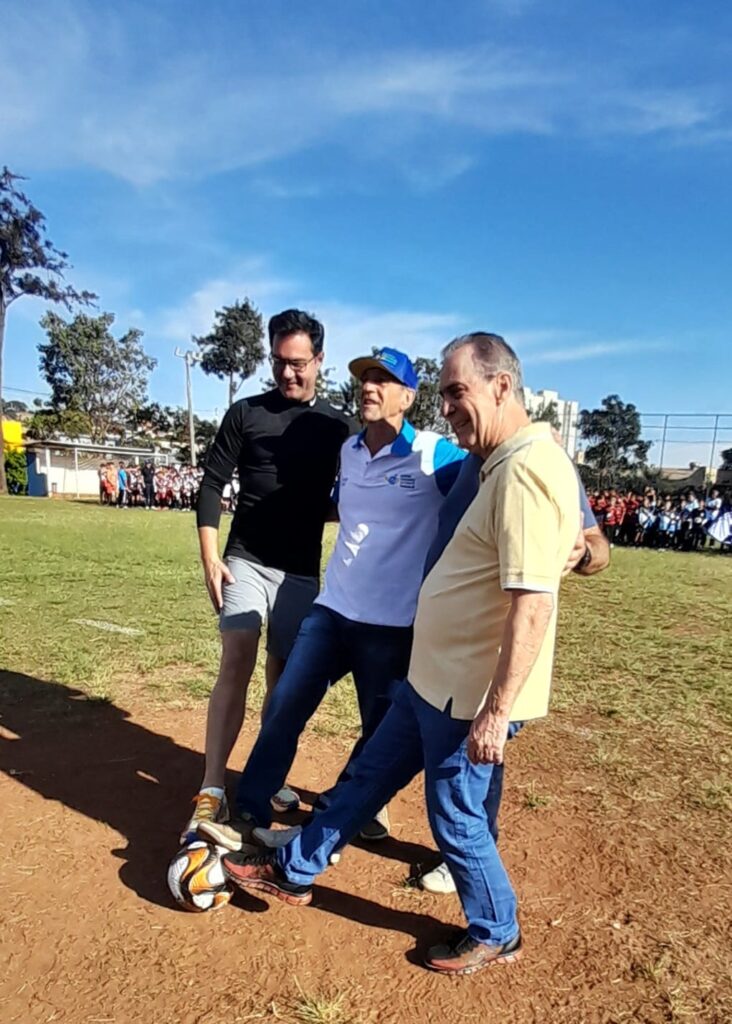 A imagem mostra três homens em um campo gramado sob um céu azul claro. Eles estão posicionados em torno de uma bola de futebol, com os pés próximos a ela, sugerindo que estão prestes a dar o pontapé inicial em um jogo ou evento.O homem à esquerda, vestindo uma camisa preta de manga comprida e shorts cinza, sorri enquanto olha para a bola. O homem do meio usa uma camisa polo branca e azul com um boné azul, e parece estar falando ou interagindo com os outros. O homem à direita, vestindo uma camisa polo amarela, também sorri e tem um braço em volta do ombro do homem do meio.Ao fundo, há uma quadra de esportes com pessoas, possivelmente crianças ou estudantes, alinhadas em uma formação. Há também árvores e edifícios visíveis ao longe, indicando um ambiente escolar ou comunitário. A iluminação sugere que a foto foi tirada durante o dia, com o sol brilhando.