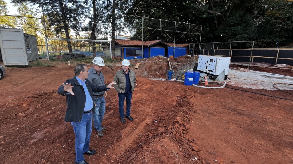 A imagem mostra três homens em um canteiro de obras. Eles estão vestindo capacetes e roupas de trabalho. O homem mais à esquerda está falando e gesticulando com as mãos. Os outros dois homens estão ouvindo. Ao fundo, há um prédio azul com telhado marrom, cercado por uma cerca de arame. Há também um gerador branco, dois barris azuis e um cone laranja. O chão é de terra vermelha, com algumas pedras e marcas de pneus. As árvores estão ao redor do local, e o céu está parcialmente nublado.