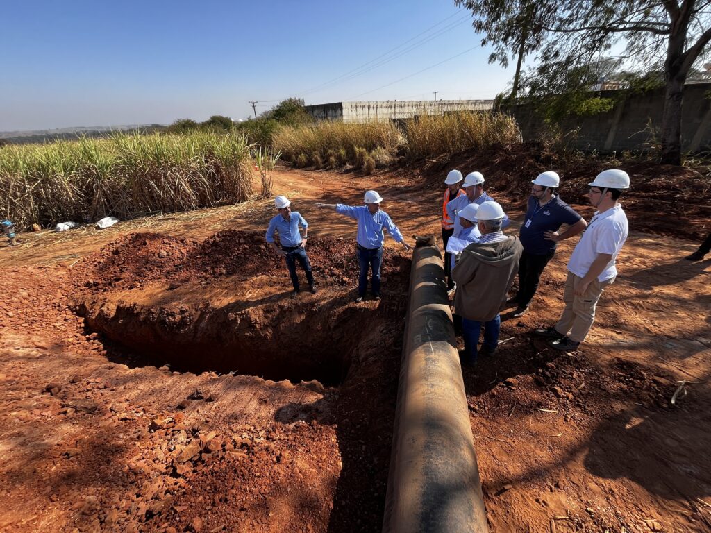 Um grupo de engenheiros está reunido em um canteiro de obras, inspecionando um grande cano exposto em uma vala. O solo ao redor é vermelho e solto, indicando uma área de construção. Ao fundo, há um campo de cana-de-açúcar e uma estrutura de concreto. O céu está azul e claro, sugerindo um dia ensolarado. Os engenheiros, todos usando capacetes brancos, parecem estar discutindo o projeto. Um deles está apontando para o cano, explicando algo aos outros. Outro engenheiro segura alguns papéis, possivelmente planos ou relatórios. A cena transmite uma sensação de trabalho em equipe e análise técnica em um ambiente externo.