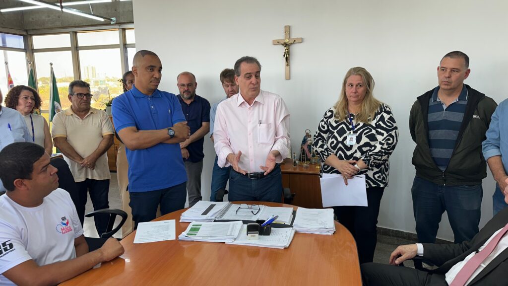 Prefeito Helinho Zanatta, de camisa rosa, fala em pé ao redor da mesa de reuniões, ladeado por lideranças comunitárias e secretários. Todos observam atentamente a fala, com documentos espalhados sobre a mesa.