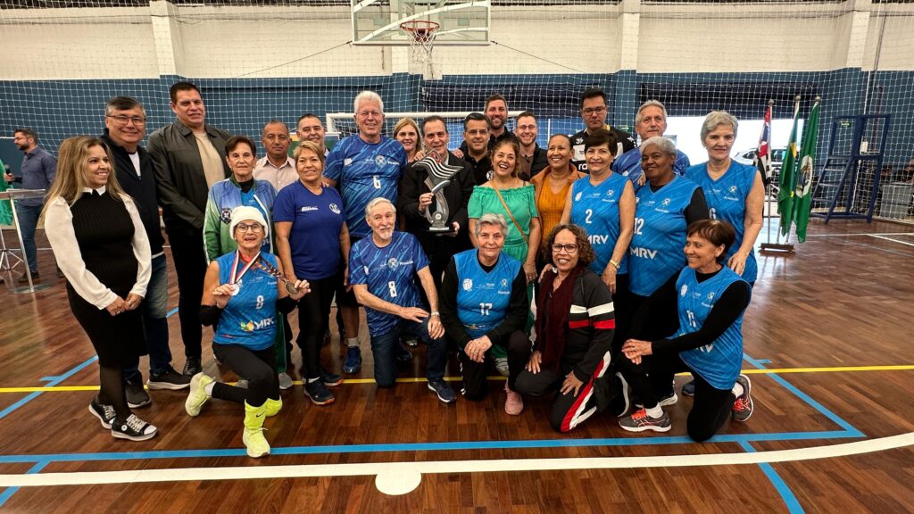 A imagem mostra um grupo de pessoas, provavelmente uma equipe esportiva, posando para uma foto em uma quadra esportiva. Há um troféu no centro da imagem, segurado por um homem. A maioria das pessoas está vestida com uniformes esportivos azuis e pretos. Algumas pessoas estão usando medalhas, o que sugere que eles podem ter vencido uma competição. O ambiente parece ser um ginásio, com uma quadra de madeira, rede de vôlei e arquibancadas ao fundo. Há também algumas bandeiras em um dos lados da foto. O grupo parece estar celebrando uma vitória ou um evento esportivo.