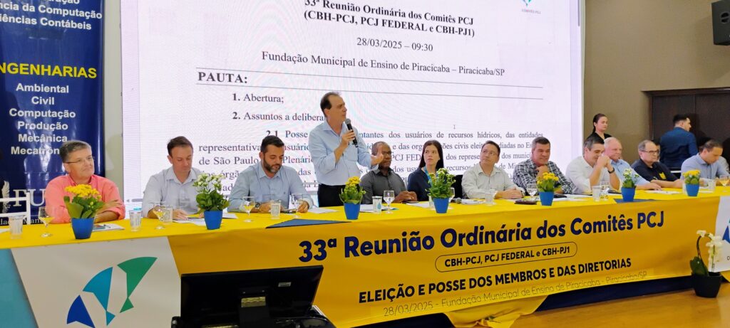 A imagem mostra um evento realizado em 28 de março de 2025, na Fundação Municipal de Ensino de Piracicaba, em Piracicaba/SP. O evento é a 33ª Reunião Ordinária dos Comitês PCJ (CBH-PCJ, PCJ FEDERAL e CBH-PJ1), com foco na eleição e posse dos membros e das diretorias. No painel principal, a pauta da reunião inclui a abertura e assuntos a serem deliberados, como a posse de representantes de usuários de recursos hídricos, entidades e órgãos civis eleitos. Na mesa principal, várias pessoas estão sentadas, algumas participando ativamente do evento, como um homem discursando ao microfone. Há também arranjos de flores sobre a mesa, que é coberta por uma faixa amarela com as informações do evento. Ao fundo, outros participantes e pessoas em pé indicam um evento com boa presença. À esquerda, um banner azul exibe os cursos oferecidos pela instituição, incluindo Engenharias (Ambiental, Civil, Computação, Produção, Mecânica, Mecatrônica).