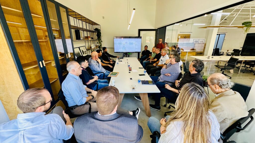 A imagem mostra um grupo de homens sentados em uma sala de reuniões moderna, com uma longa mesa branca no centro. Alguns homens estão olhando para a câmera, enquanto outros parecem estar conversando ou prestando atenção a algo fora do quadro. A sala é iluminada por luz natural que entra pelas grandes janelas, e há prateleiras com livros e decorações nas paredes. Um laptop está aberto sobre a mesa, sugerindo que uma apresentação ou reunião de trabalho está em andamento. A atmosfera geral parece profissional e colaborativa.