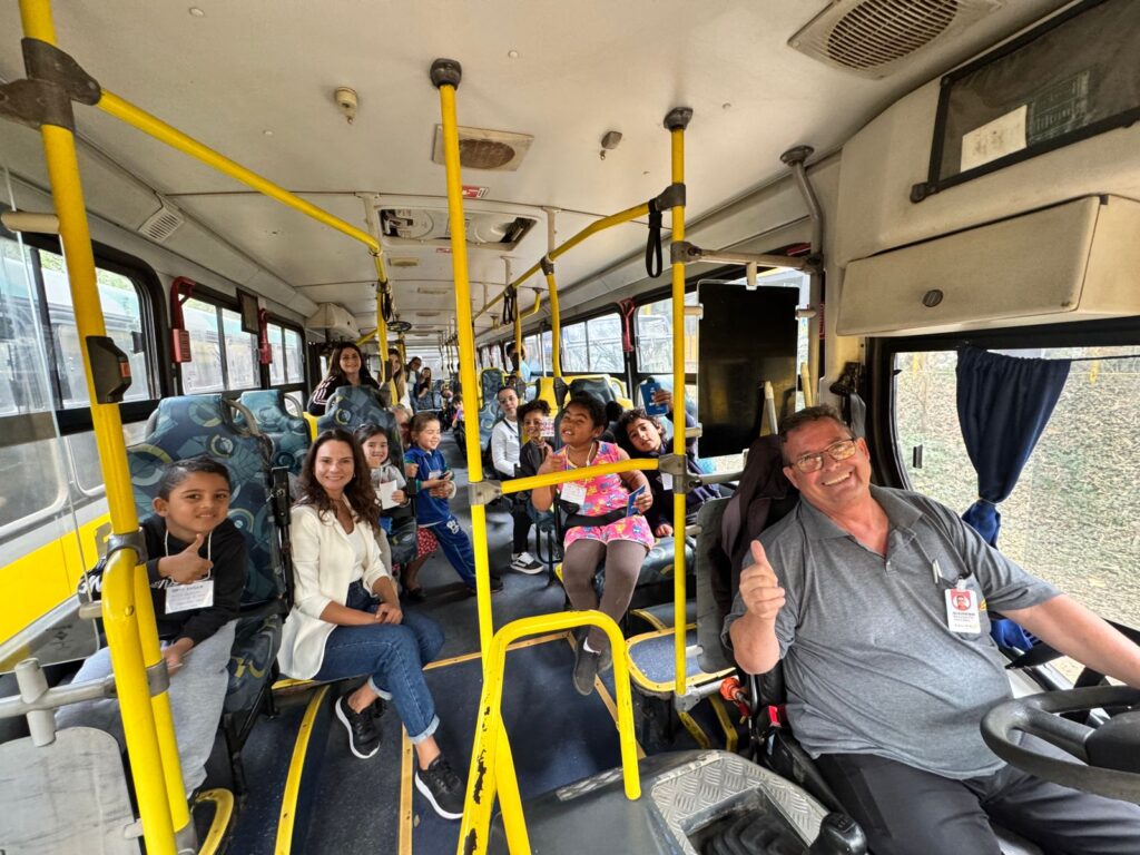 Crianças e uma professora sorriem dentro de um ônibus escolar amarelo. No banco da frente, o motorista sorri e faz sinal de positivo para a foto.