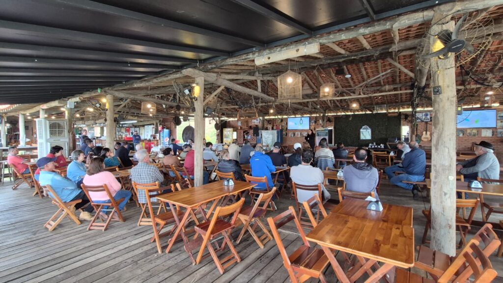 A imagem retrata um evento comunitário ou reunião em um espaço rústico e acolhedor, com piso e teto de madeira. As pessoas estão sentadas em cadeiras de madeira, voltadas para uma área de apresentação onde um palestrante se posiciona ao lado de uma tela grande. O ambiente é aberto, com iluminação natural e artificial, incluindo luminárias pendentes e ventiladores. Há elementos decorativos que dão um toque informal e cultural ao local, como um violão pendurado na parede, um ventilador grande e uma máquina de refrigerante da Pepsi.