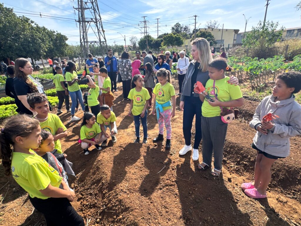 🌿 A imagem retrata um momento educativo em uma horta comunitária, onde crianças com camisetas amarelo-limão interagem com adultos, aprendendo sobre o cultivo de plantas. Algumas seguram mudas ou flores, enquanto ao fundo aparecem canteiros verdes, árvores e estruturas urbanas como postes e fios elétricos.