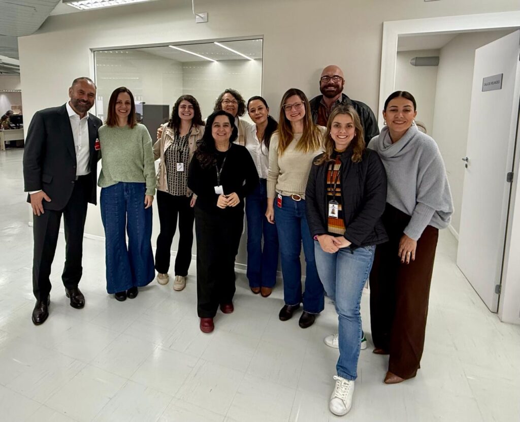 Essa imagem mostra um grupo de nove pessoas reunidas em um ambiente profissional, possivelmente um escritório ou local de trabalho. Estão vestidas com roupas casuais de trabalho, e algumas usam crachás de identificação. Ao fundo, há móveis de escritório e uma porta com a placa “Auditório”.