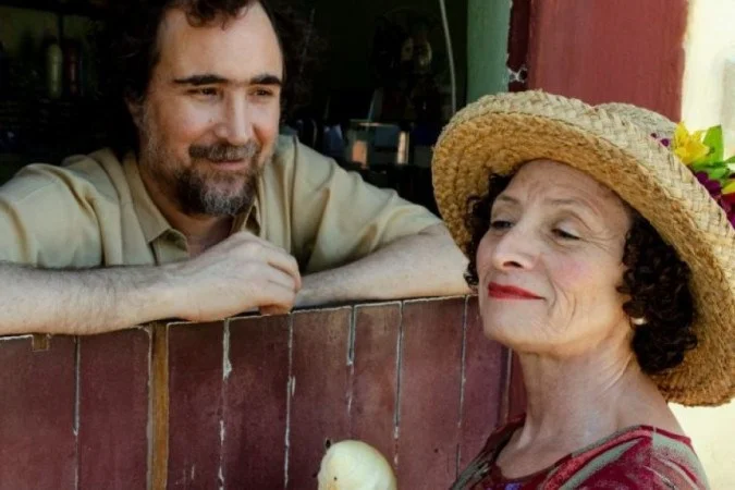 Cena em que uma mulher, com chapéu de palha enfeitado, olha sorridente e de perfil, enquanto um homem barbudo, encostado em uma janela de madeira, observa a personagem com olhar afetuoso.