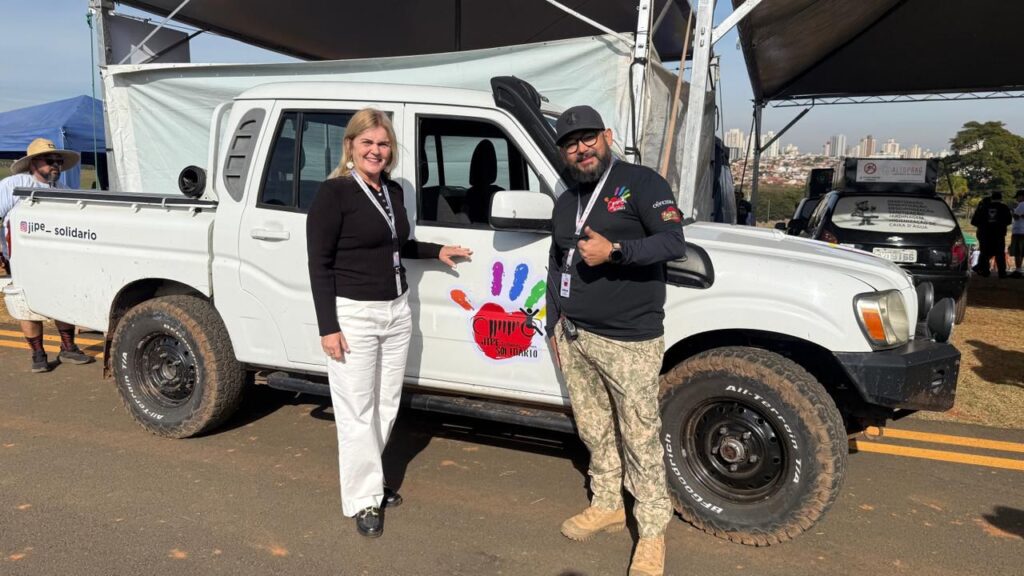 Mulher e homem posam sorrindo ao lado de caminhonete branca com logotipo do “Jipe Solidário”; o homem faz sinal de positivo.