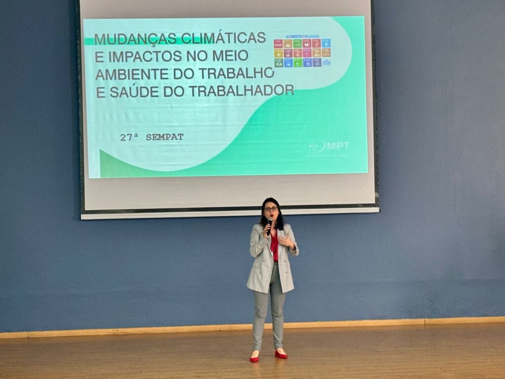 A imagem mostra uma apresentação sendo realizada em um auditório, com uma pessoa em pé no palco diante de uma tela grande que exibe um slide temático. O ambiente é formal e voltado à discussão de questões relacionadas ao meio ambiente do trabalho e à saúde dos trabalhadores.