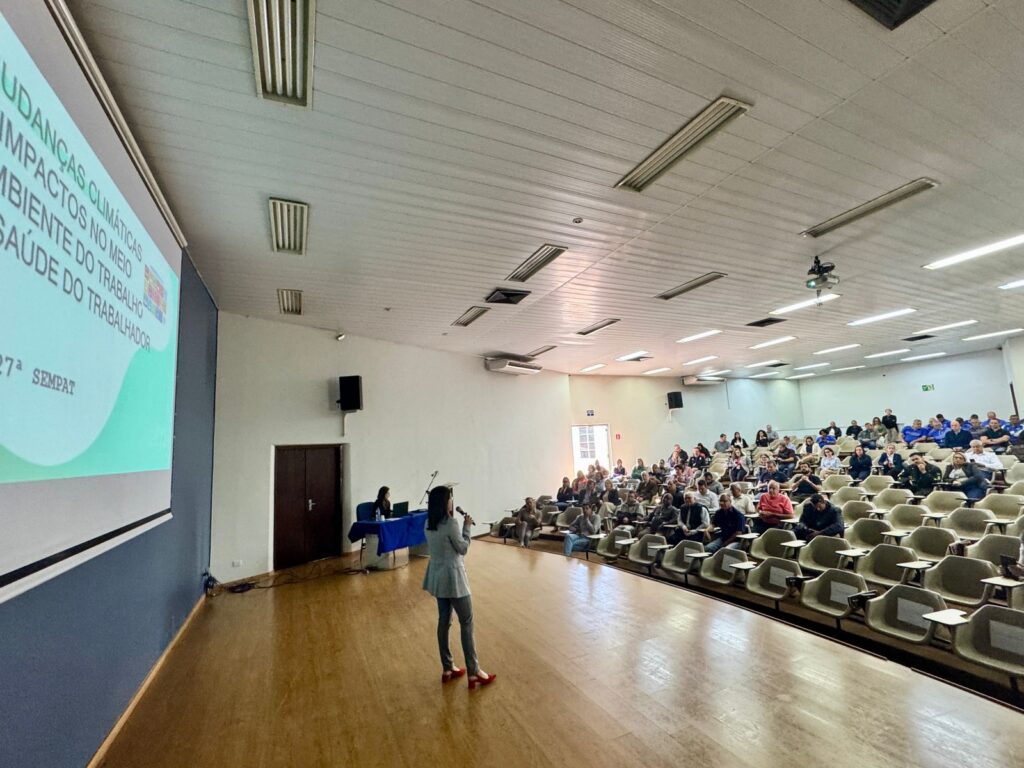 A imagem retrata uma palestra em andamento em um auditório amplo e bem iluminado. Uma pessoa está em pé no palco, falando ao microfone diante de uma plateia atenta, enquanto outra permanece sentada à mesa, possivelmente como mediadora ou organizadora. O público está distribuído em fileiras de cadeiras, demonstrando interesse pelo conteúdo apresentado. A atmosfera é formal e voltada ao compartilhamento de conhecimento, sugerindo um evento profissional voltado a temas relevantes para o ambiente de trabalho e a saúde.