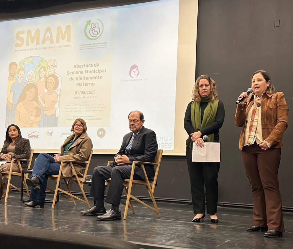 A imagem mostra um evento de abertura da Semana Municipal de Aleitamento Materno. No centro da imagem, há uma tela com o título do evento, data, horário, local e link para inscrição. Cinco pessoas estão sentadas em cadeiras no palco, e duas mulheres estão em pé, uma delas falando ao microfone. As pessoas sentadas são: uma mulher à esquerda, duas mulheres no centro e um homem à direita. As duas mulheres em pé estão à direita, uma delas segurando um papel e a outra falando ao microfone. A iluminação é suave e o fundo é escuro, com a tela iluminada ao fundo.