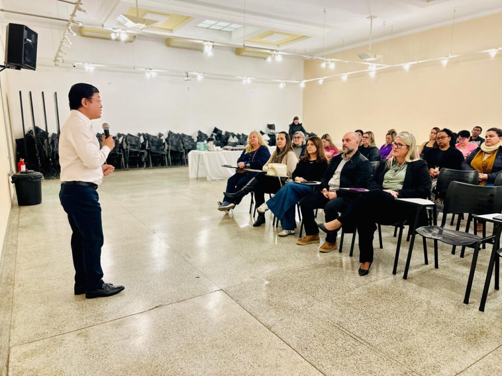 A imagem mostra um evento formal, como uma palestra ou seminário. Uma pessoa está em pé, falando ao microfone diante de uma plateia sentada em cadeiras organizadas em fileiras. O ambiente é bem iluminado, com teto branco e piso polido, transmitindo uma atmosfera profissional. Ao fundo, há cadeiras empilhadas e uma mesa com lanches ou bebidas, sugerindo que o evento oferece algum tipo de recepção.