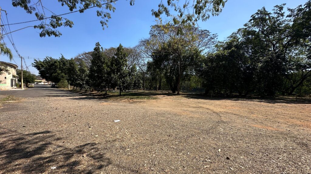 A imagem mostra uma área externa em dia ensolarado, com céu limpo e azul. Em primeiro plano, há um terreno de terra e cascalho, aparentemente preparado para obras ou intervenções futuras. Ao fundo, vê-se uma estrada pavimentada, algumas edificações e postes com fiação elétrica, sugerindo proximidade com uma zona urbana ou industrial. A vegetação ao redor é abundante, com árvores e arbustos que delimitam o espaço.