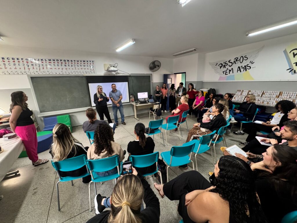 A imagem mostra uma sala de aula organizada para uma atividade formativa, onde um grupo de adultos está sentado em semicírculo, atentos à apresentação de duas pessoas que falam na frente da sala. O ambiente é acolhedor e educativo, com cartazes coloridos nas paredes, incluindo um que diz “PÁSSAROS TÊM ASAS” e outro com a palavra “VOAR” acompanhada de um desenho estilizado de uma ave. Há também um quadro negro, um computador sobre a mesa e materiais pedagógicos espalhados pelo espaço.