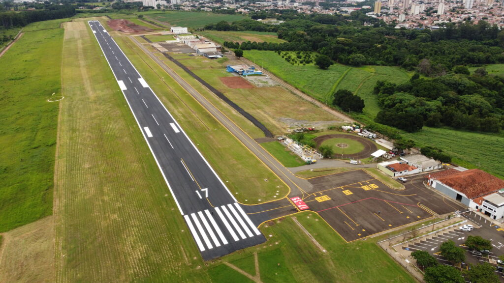 A imagem mostra uma vista aérea de um pequeno aeroporto cercado por áreas verdes e desenvolvimento urbano. O destaque é uma pista de asfalto com a marcação “17” em uma das extremidades, indicando sua orientação magnética. Ao lado da pista, há taxiways com sinalização amarela que levam a uma área de pátio com hangares, edifícios operacionais e veículos estacionados. Também é visível um heliponto circular próximo às construções e um estacionamento com diversos carros. Ao redor, há campos gramados, árvores e uma zona urbana densa ao fundo.