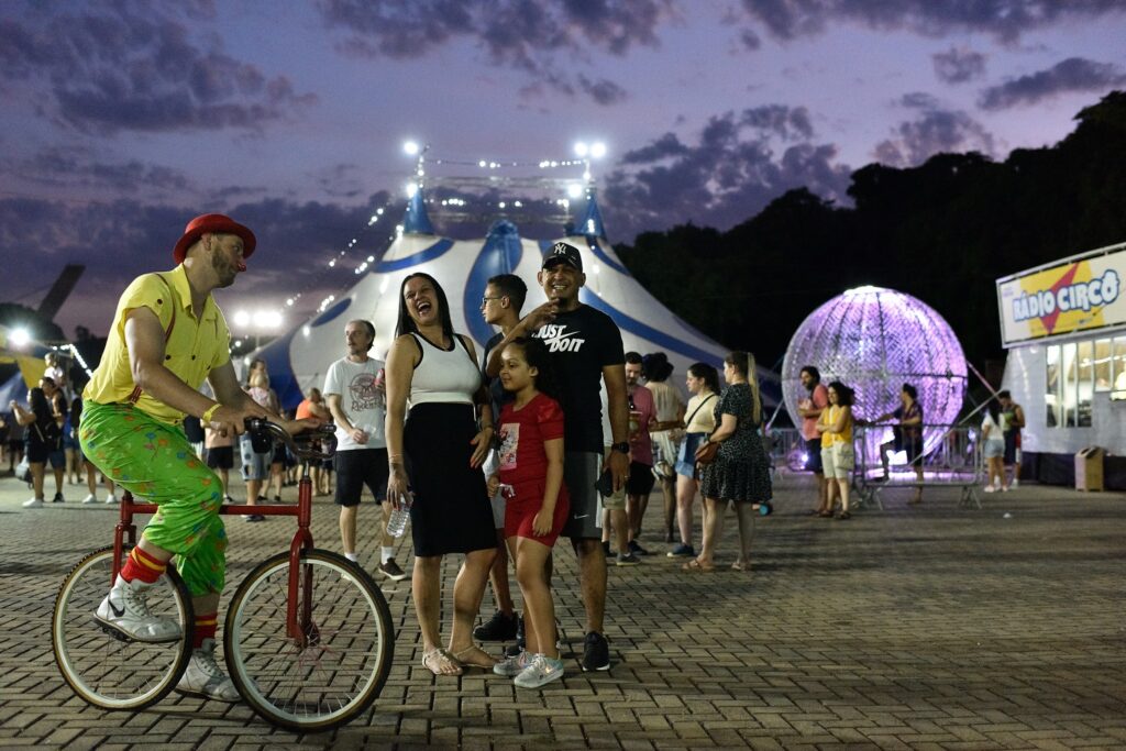 Área externa do circo à noite: famílias e grupos de amigos circulam pelo espaço. Um palhaço em bicicleta interage com o público, enquanto ao fundo a esfera metálica iluminada e a lona do circo criam um cenário vibrante e acolhedor.