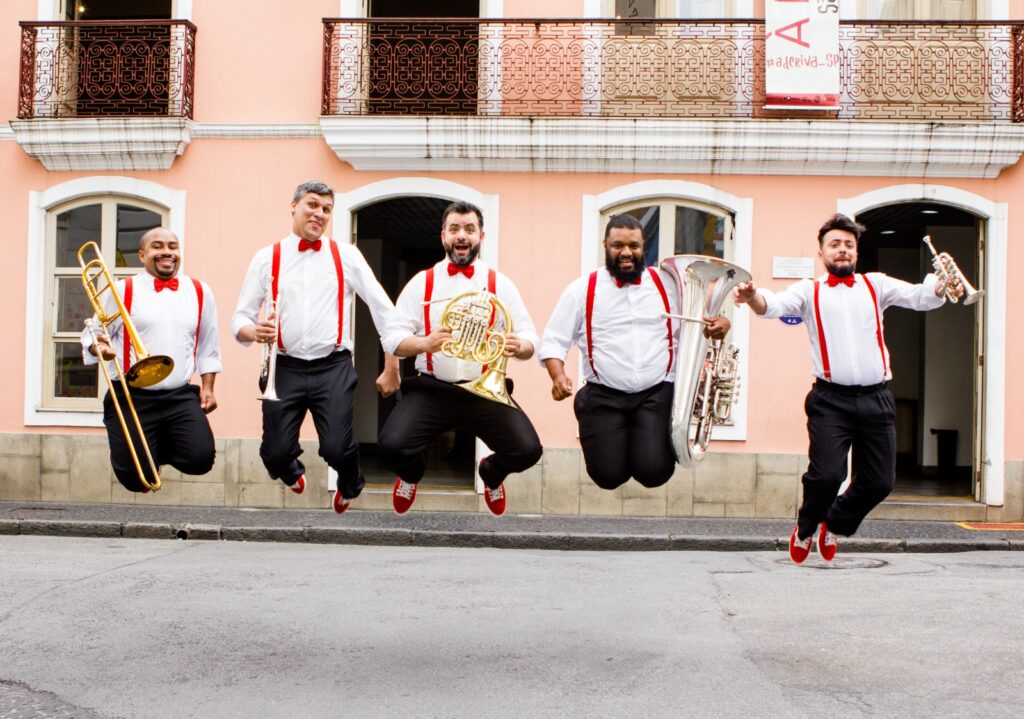 Cinco músicos do grupo BrassUka estão no ar, pulando em sincronia, com expressão alegre. Usam camisas brancas, calças pretas, sapatos vermelhos, gravatas-borboleta vermelhas e suspensórios vermelhos. Cada um segura seu instrumento de sopro: trombone, trompete, trompa, tuba e flugelhorn. Ao fundo, a fachada de um prédio antigo de cor rosa com janelas e sacadas de ferro trabalhado.