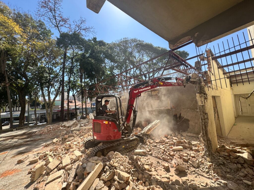 Pessoa opera uma escavadeira vermelha derrubando paredes de um prédio, com entulho espalhado no chão e árvores ao fundo em um dia ensolarado.