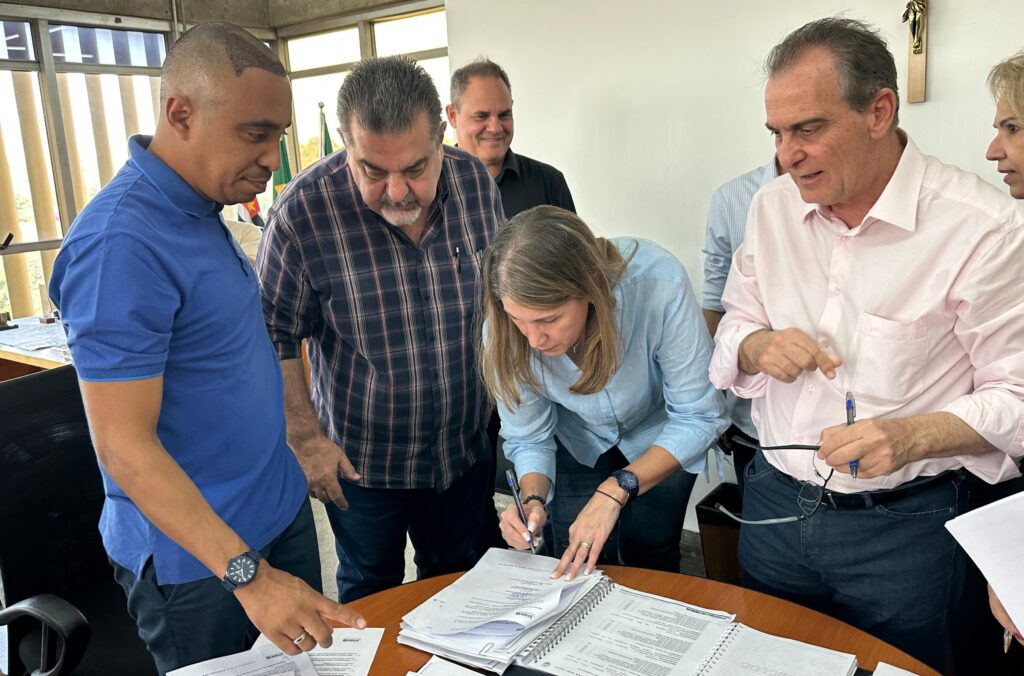 Mulher de camisa azul, representante da Orquestra Sinfônica de Piracicaba, assina o termo sobre a mesa, acompanhada pelo prefeito e secretários. Os demais participantes acompanham a assinatura sorrindo.