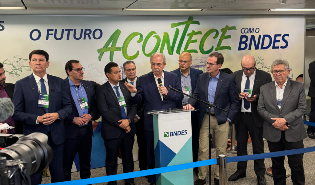 A imagem mostra um grupo de homens em um evento, em frente a um banner com a frase "O FUTURO ACONTECE COM O BNDES". Há um homem em destaque, falando ao microfone em um púlpito com o logo do BNDES. Os outros homens estão ao redor, alguns olhando para o orador, outros para a frente. Todos parecem estar vestindo ternos ou blazers e crachás. Há também equipamentos de mídia, como câmeras, na frente.