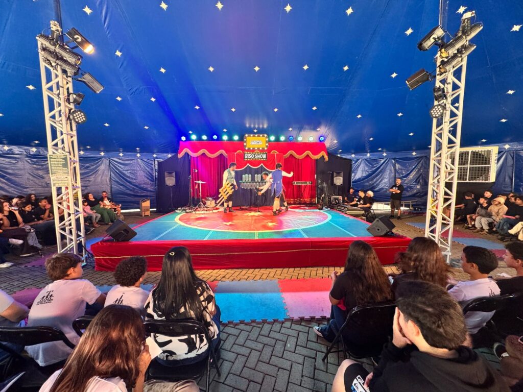 Cena dentro de uma lona de circo, com público de jovens assistindo atentos a um artista que se apresenta no picadeiro circular. O artista está de pé, equilibrando objetos, sob iluminação colorida.