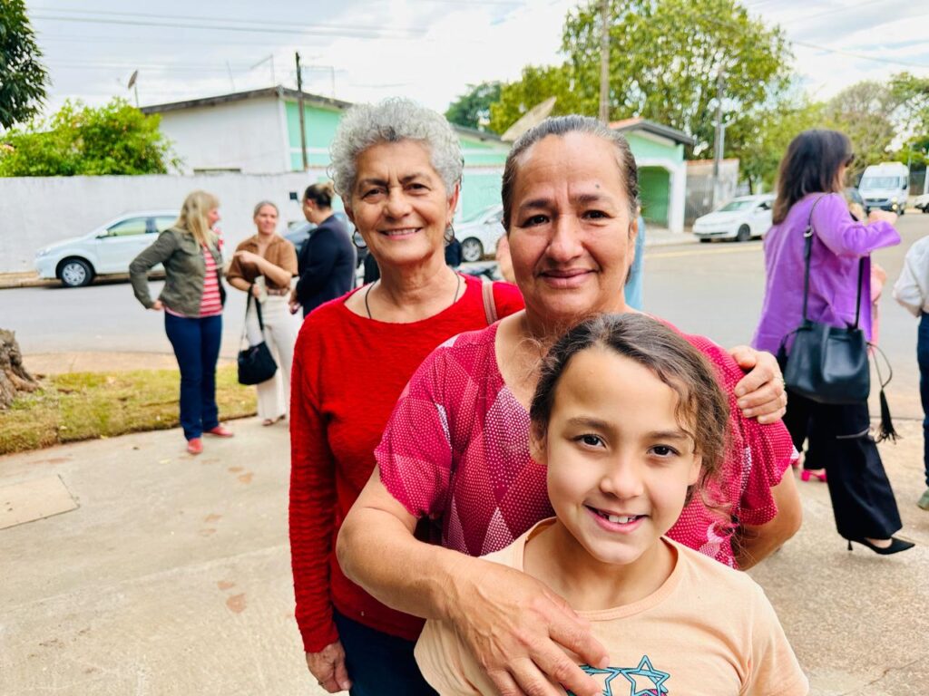 A imagem mostra três mulheres em primeiro plano, sorrindo para a câmera. A mulher mais velha, à esquerda, tem cabelos grisalhos curtos e usa uma blusa vermelha. Ao lado dela, uma mulher de meia-idade com cabelos escuros e lisos usa uma blusa rosa com um padrão. No centro, uma menina com cabelos castanhos ondulados e um sorriso largo usa uma camiseta de cor pêssego.