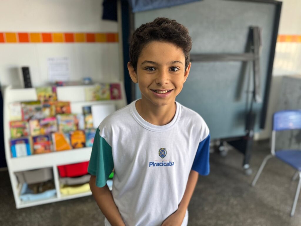 A imagem mostra uma pessoa vestindo uma camiseta branca com detalhes azuis, estampada com a palavra “Piracicaba” e um logotipo, sugerindo vínculo com alguma instituição local. Ao fundo, há uma estante repleta de livros e objetos coloridos, uma cadeira azul e um quadro negro, compondo um ambiente que remete a uma sala de aula ou espaço educativo.