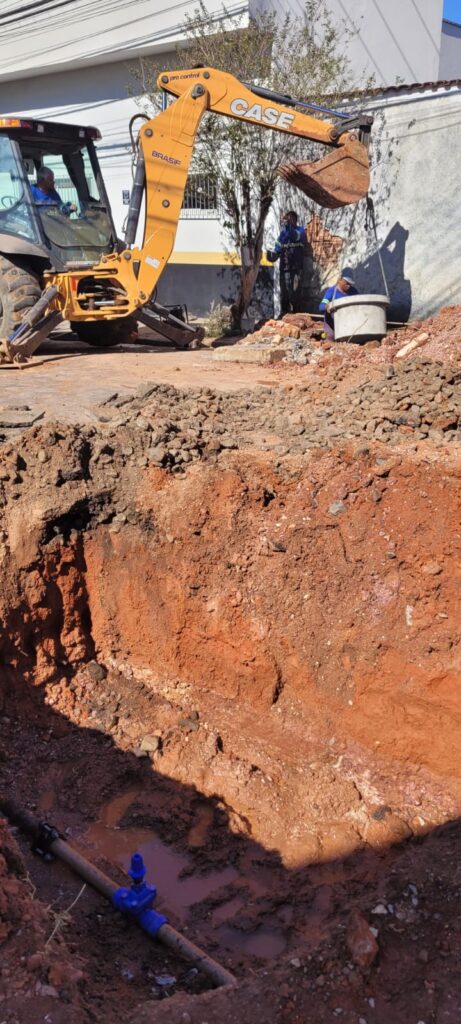 A imagem mostra uma escavadeira amarela da marca CASE operando em uma obra. Um trabalhador está dentro da cabine da escavadeira, e outros dois trabalhadores estão do lado de fora, próximos à máquina. O foco principal da imagem é uma vala recém-escavada, com terra avermelhada exposta nas paredes. No fundo da vala, é possível ver um cano com uma válvula azul, indicando que se trata de uma obra de infraestrutura, possivelmente relacionada ao abastecimento de água ou saneamento. Há também água acumulada no fundo da vala, o que é comum em escavações. Ao fundo, observa-se uma construção com paredes claras e algumas árvores. A cena sugere um dia de trabalho intenso em uma área urbana ou suburbana.
