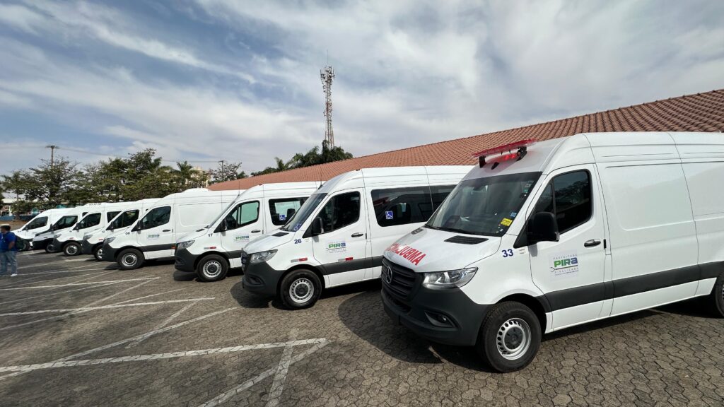 A imagem mostra uma fila de ambulâncias brancas estacionadas em um pátio com piso de paralelepípedos. As ambulâncias são modernas e parecem ser do mesmo modelo, com portas deslizantes e janelas. Várias delas exibem o logotipo "PREFEITURA PIRA" e números de identificação, como "26" e "33". Uma das ambulâncias na frente tem uma barra de luzes vermelhas e azuis no teto e a palavra "AMBULÂNCIA" escrita em vermelho no capô. Ao fundo, há uma construção com telhado de telhas marrons e uma torre de telefonia celular. O céu está parcialmente nublado.