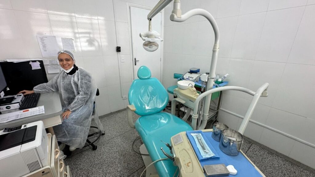 A imagem mostra um consultório odontológico com uma profissional sentada em frente a um computador. Ela está usando um jaleco descartável e uma touca. Ao lado dela, há uma cadeira odontológica azul, um equipo odontológico com instrumentos e uma impressora. O consultório parece limpo e organizado, com paredes revestidas de azulejos brancos.